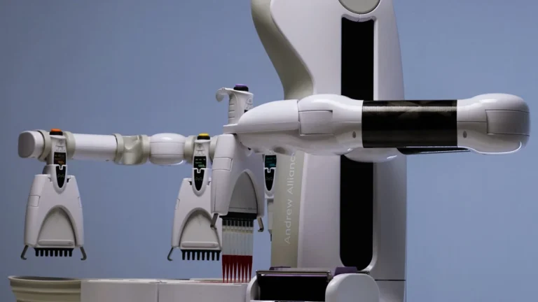 Biały robot laboratoryjny z ramieniem trzymającym wielokanałową pipetę nad statywem, obok kilka pipet elektronicznych na tle niebieskiej ściany